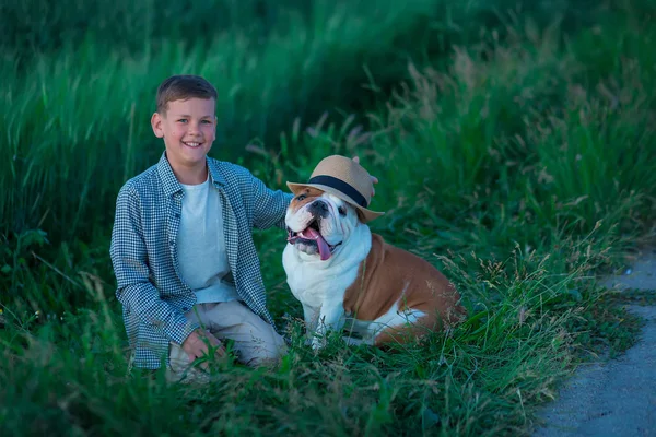 小男孩和他的英国公牛狗站在绿色黑麦的草地上 Handsom 孩子摆姿势与最好的朋友穿着时尚的衣服和质朴复古的帽子 — 图库照片