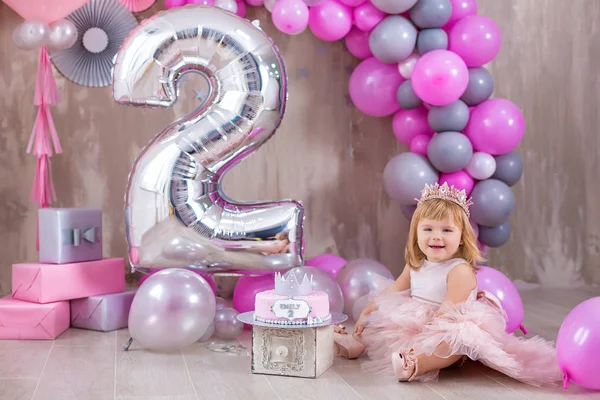 Princess baby girl celebrating life event wearing golden crown and pink airy dress. Cute girl posing in pastel colors studio shoot with air baloons and cake