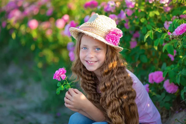 Şirin esmer çocuk mavi gözlü kız holding ayakta pembe gül alanındaki çiçekler. Şık elbise ve şapka giymiş, yaz saati gün çocukluk zevk.
