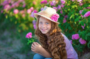 Şirin esmer çocuk mavi gözlü kız holding ayakta pembe gül alanındaki çiçekler. Şık elbise ve şapka giymiş, yaz saati gün çocukluk zevk.