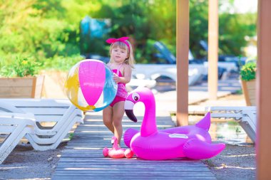 Bebek kız pembe flamingo ile sahilde ahşap zemin üzerinde poz şık yüzme giyim giymiş. Olay yerinde tatil gülümseyen bir sevimli bayan deniz tarafında ve hayattan zevk zaman çocukluk bebeklik