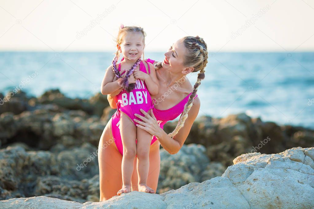 Mother daughter having fun resting on the rocky beach wearing pink swimming suits. Blond lady with girl enjoy summer time together Swimming suits deisigned by photographer.