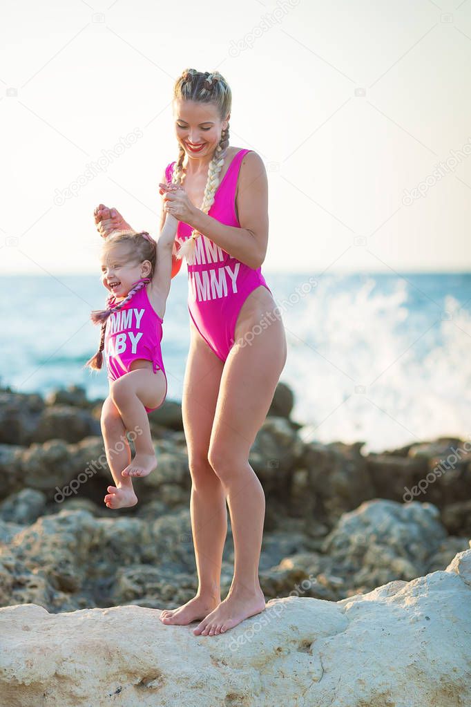Mother daughter having fun resting on the rocky beach wearing pink swimming suits. Blond lady with girl enjoy summer time together Swimming suits deisigned by photographer.