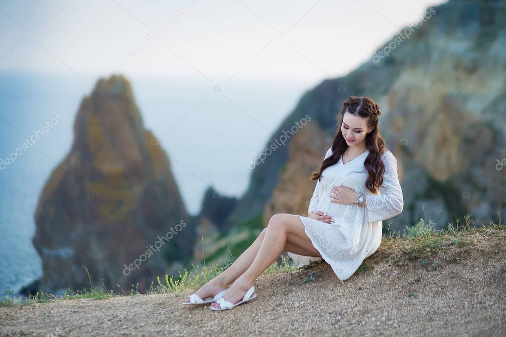 Pregnant woman posing sitting on cliff mountain wearing airy whit dress with blue sea on background.