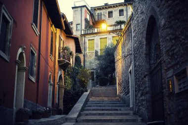 Verona, İtalya 'da merdivenleri olan dar bir sokak. Kırmızı ve turuncu binalar ve ahşap pencereler. Eski mimarisi ve yeşil çiçekleri olan bir şehir caddesi. Akşam gizemli ışıklar.