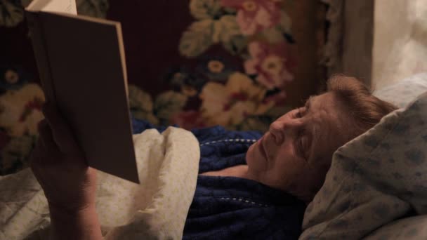 Una anciana anciana está acostada en la cama bajo una manta y leyendo un libro — Vídeos de Stock