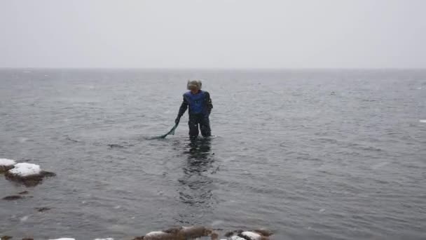 Pescador con una red de pesca camina a lo largo de la playa en invierno y recoge delicias — Vídeos de Stock