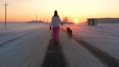 Hareket, kış akşam soğuk havalarda bir romantik kırmızı günbatımı, karla kaplı alanların etrafında arka planı yolda köpeğiyle yürüyen kadın arkadan görünüm. Ağır çekim, 4k.