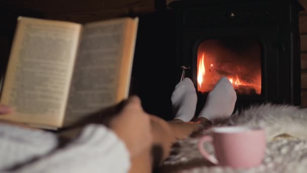 Женщина лежит на мягком ковре перед камином и читает книгу — стоковое видео