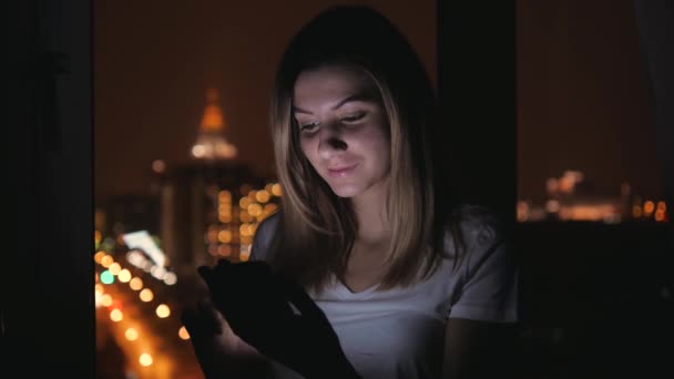 Увечері жінка стоїть біля вікна, вдома використовує телефон додатків — стокове відео
