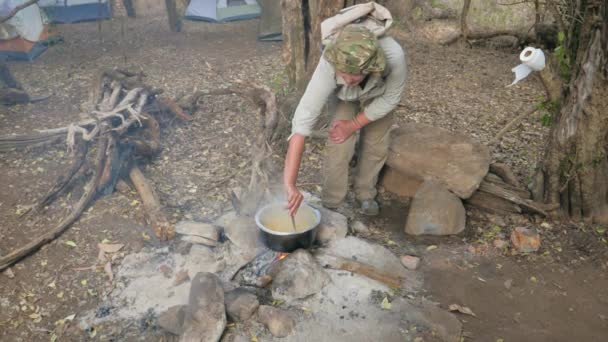 Turista en el camping agitado con una cuchara para cocinar pasta en una cacerola en el fuego — Vídeo de stock