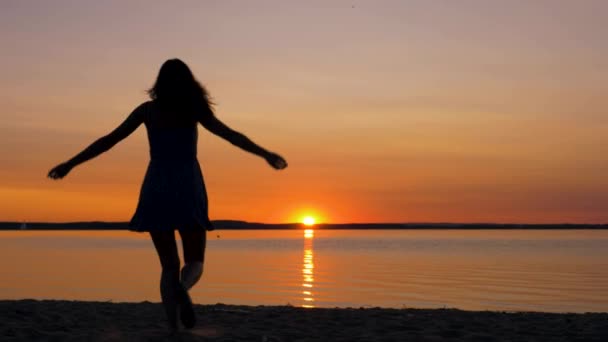 Жінка проходить по відношенню до заходу сонця на пляжі, піднімаючи руки вгору і обертаючись — стокове відео