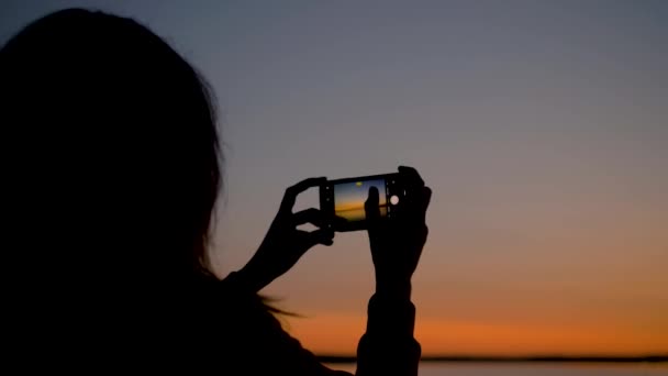 女性は海の海岸に夕日のスマート フォンで写真を撮る — ストック動画