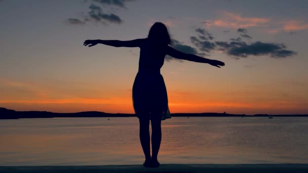 Sziluettje egy nő emelte a kezét az oldalára a vízparton a naplemente — Stock videók
