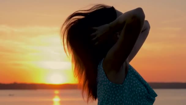 На пляже красивая женщина в платье, сидя на золотом закате исправляет волосы — стоковое видео