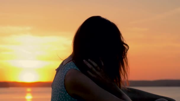 Na praia, a mulher sentada ao pôr do sol dourado corrige e acaricia seu cabelo — Vídeo de Stock