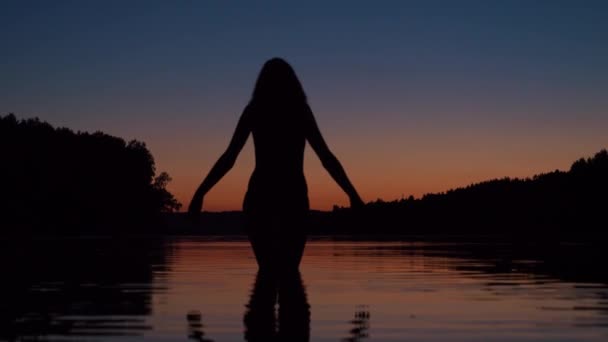 Mujer de pie hasta la rodilla en el agua del lago al atardecer levanta sus brazos — Vídeos de Stock