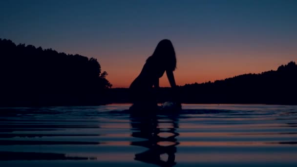 女性のシルエットは、膝を立っている夕日およびはねかける水で湖の奥深くに — ストック動画