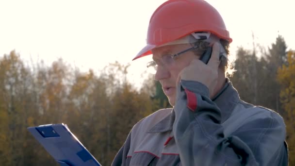 Construtor em um capacete conversando no telefone olhando para o plano contra o sol — Vídeo de Stock