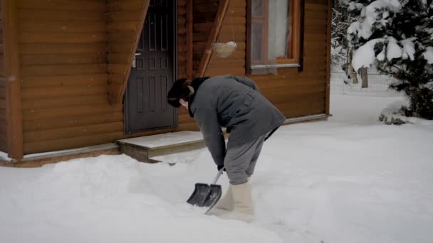 El hombre limpia un patio de palas de la nieve en el fondo de una casa de madera en invierno — Vídeo de stock