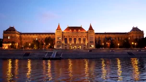 从多瑙河上的多瑙河上的布达佩斯技术和经济大学 在许多汽车驾驶的海滨 在晚上与在温暖的灯光下照亮 — 图库视频影像