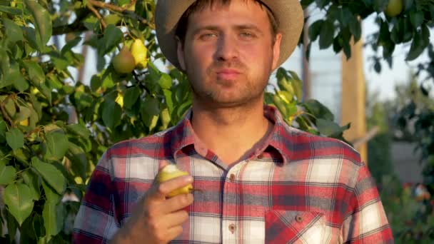 Чоловік фермер їсть грушу на фоні фруктового саду — стокове відео