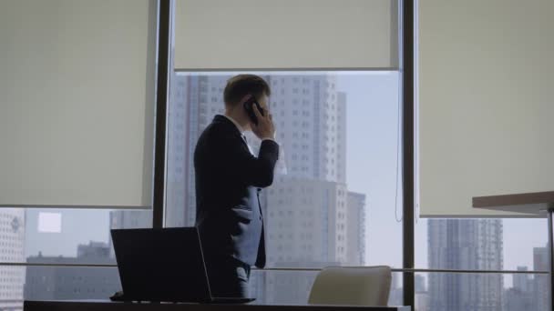Бизнесмен разговаривает по телефону в окне офиса, а затем садится на рабочее место — стоковое видео
