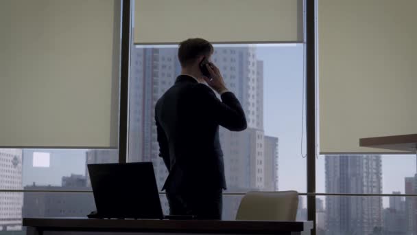 Директор в деловом костюме разговаривает по телефону в офисе окна рядом с рабочим местом — стоковое видео