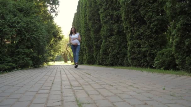 Вагітна кавказька жінка, яка ходить по алеї в парку і хитає своїм животом — стокове відео