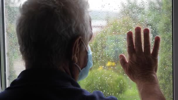 Старик в защитной медицинской маске выглядывает в окно и кладет руку на стекло — стоковое видео