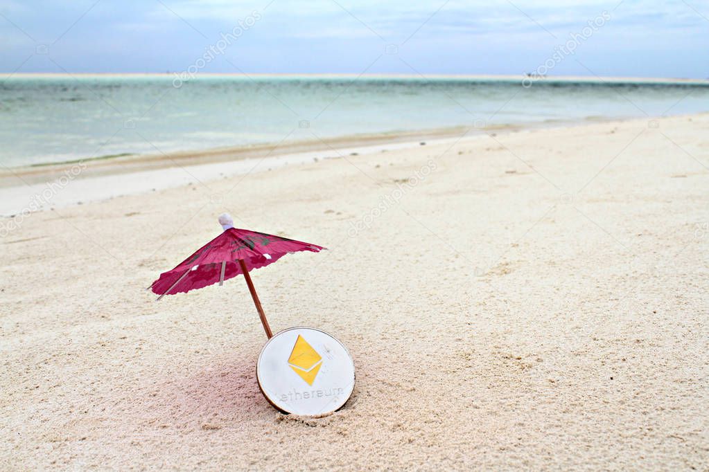 Silver Ethereum coin under the paper umbrella on the beach, shallow focus