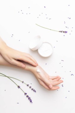 Beautiful well-conditioned female hands moisturizing cream surrounded with lavender flowers on the white background. Hand and body care concept clipart