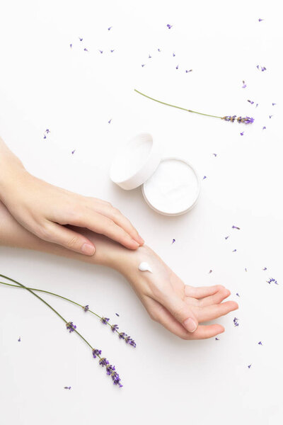 Beautiful well-conditioned female hands moisturizing cream surrounded with lavender flowers on the white background. Hand and body care concept