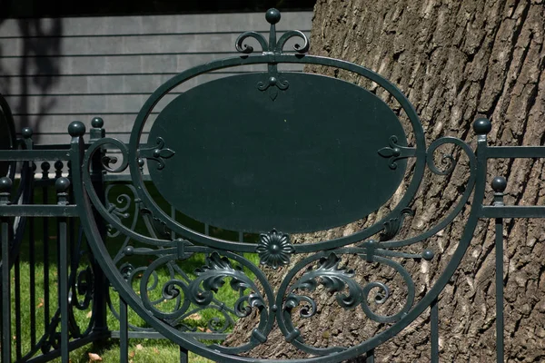 Büyük ağacın önündeki metal dövme çitinde boş boş tabela. Mo