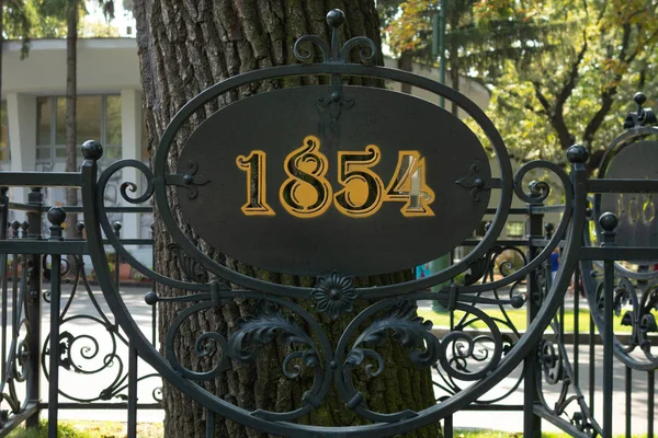 1854 year sign on metal forging fence in front of big tree. Info