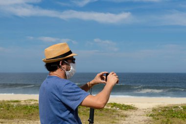 Yeni normal, orta yaşlı adam plajın bir fotoğrafını çekiyor. Korunmasız bir yerde. Koruyucu bir maske takıyor.