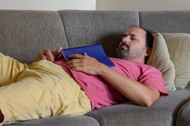 Orta yaşlı bir adam kanepede uyuyor ve karnında kitap var.