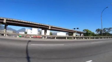 Rio de Janeiro 'daki çevre yolu modern bir binanın önünden geçen hareket halindeki bir arabanın yanından görülüyor.