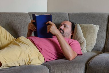 Orta yaşlı bir adam kanepede kitap okuyor.