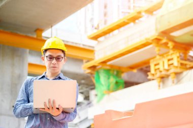 Genç Asyalı mühendisler inşaat alanında çalışıyorlar. Sarı kask emniyeti takın. Elle tutulan bilgisayar dizüstü