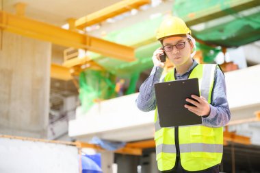 Genç Asyalı mühendisler inşaat alanında çalışıyorlar. Sarı kask ve üniforma giyin. İletişim ve evrak işleri için taşınabilir radyo alıcısı.