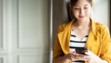 Asyalı bir kadın güzel bir gülümsemeyle ayakta duruyor ve kapalı alanda dinlenirken cep telefonuyla uygulama sohbeti yapıyor.