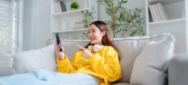 Mutlu Asyalı genç kadın kulaklık takıyor ve evdeki rahat koltukta dinleniyor, akıllı telefondan mesajlaşıyor, gülümseyen kız cep telefonu sohbetini kullanıyor, internetten alışveriş yapıyor, video görüşmesi yapıyor.