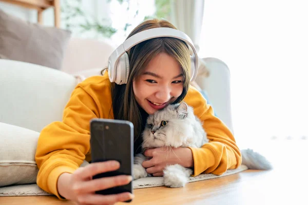 Spending your free time at home with your cat. young asian woman in headphones, with mobile phone, lies on floor in living room, with fluffy Maine Coon cat, listens to music