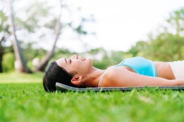 Spor elbiseli Asyalı bir kadın parkta yeşil çimlerde dinleniyor.