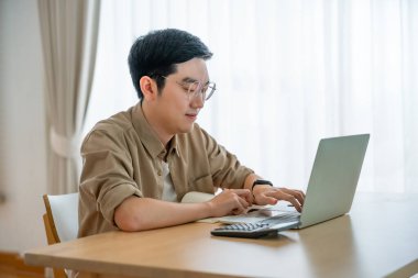 Oturma odasında dizüstü bilgisayarla çalışan mutlu Asyalı genç adam. Serbest çalışan erkek, masa başında oturmuş Webinar izlerken notlar yazıyor, internetten çalışıyor.