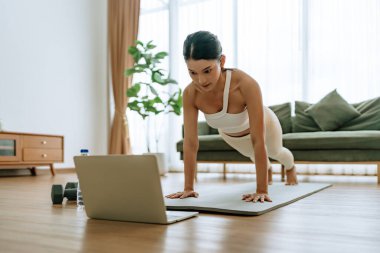 Spor kıyafetli genç bir Asyalı kadın oturma odasında dizüstü bilgisayarla antrenman dersleri izlerken antrenman minderinde tahta döşeme yapıyor. Spor ve eğlence konsepti