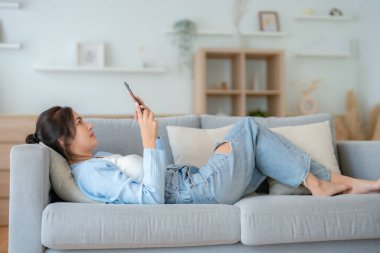 Mutlu, pozitif, genç, Asyalı bir kadın evde telefonda, gülümseyen bir kız. Uygulama kullanıyor ve internette akıllı telefonuyla kanepeye uzanıyor.