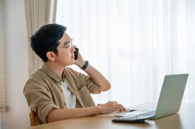 Oturma odasında dizüstü bilgisayarla çalışan mutlu Asyalı genç adam. Serbest çalışan erkek, masa başında oturmuş Webinar izlerken notlar yazıyor, internetten çalışıyor.