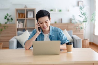 Oturma odasında dizüstü bilgisayarla çalışan mutlu Asyalı genç adam. Serbest çalışan erkek, masa başında oturmuş Webinar izlerken notlar yazıyor, internetten çalışıyor.